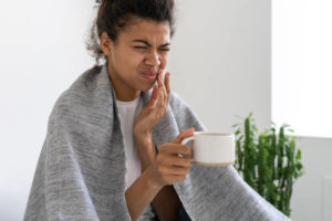 A woman wrapped in a blanket while holding a tea mug winces in pain as she touches her left jaw.